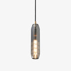 A tall Clérachic pendant with a smoke grey glass shade and a brass top, lit with a filament bulb, on a plain white backdrop.