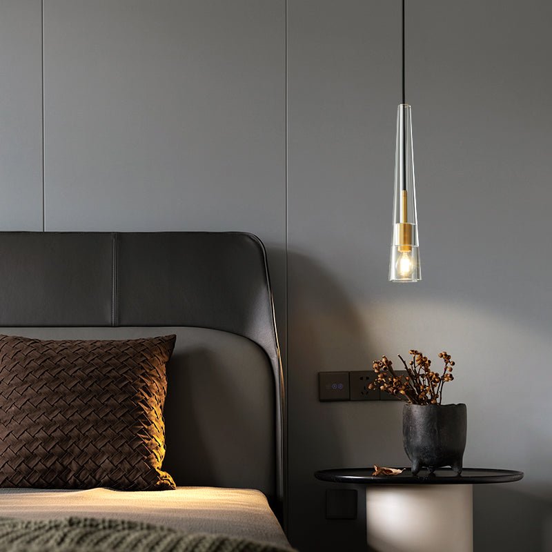 A slim, tapered clear glass Clérachic pendant with a brass collar hangs beside a leather headboard and a dark side table.
