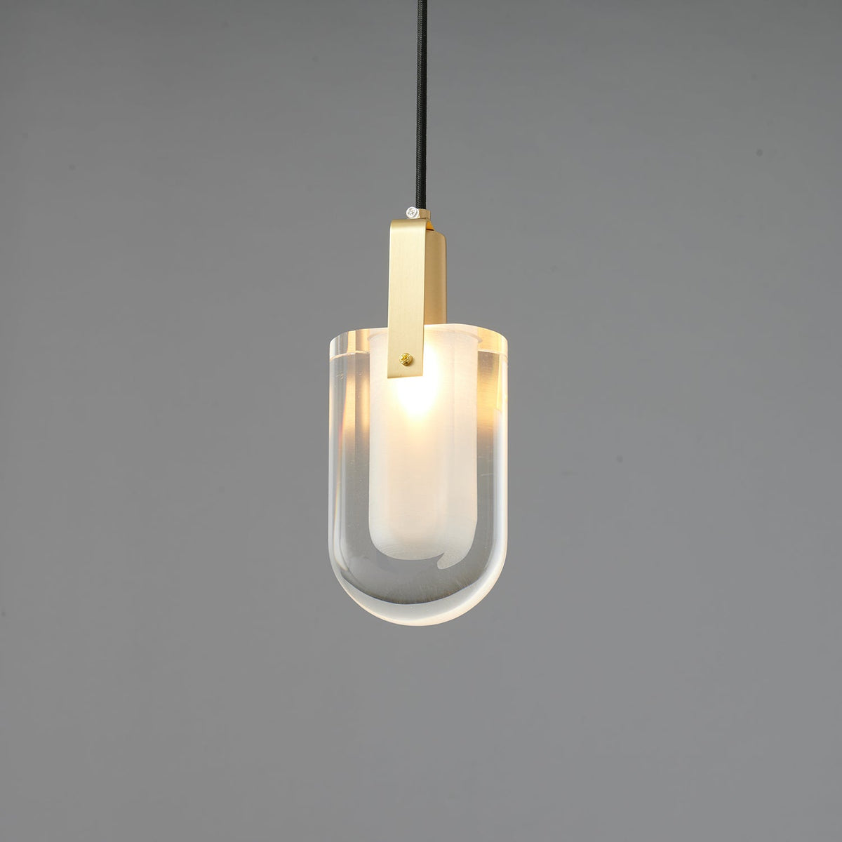 A smaller view of the lit U-shaped Clérachic pendant, with a brass bracket and a clear glass outer shade on grey.