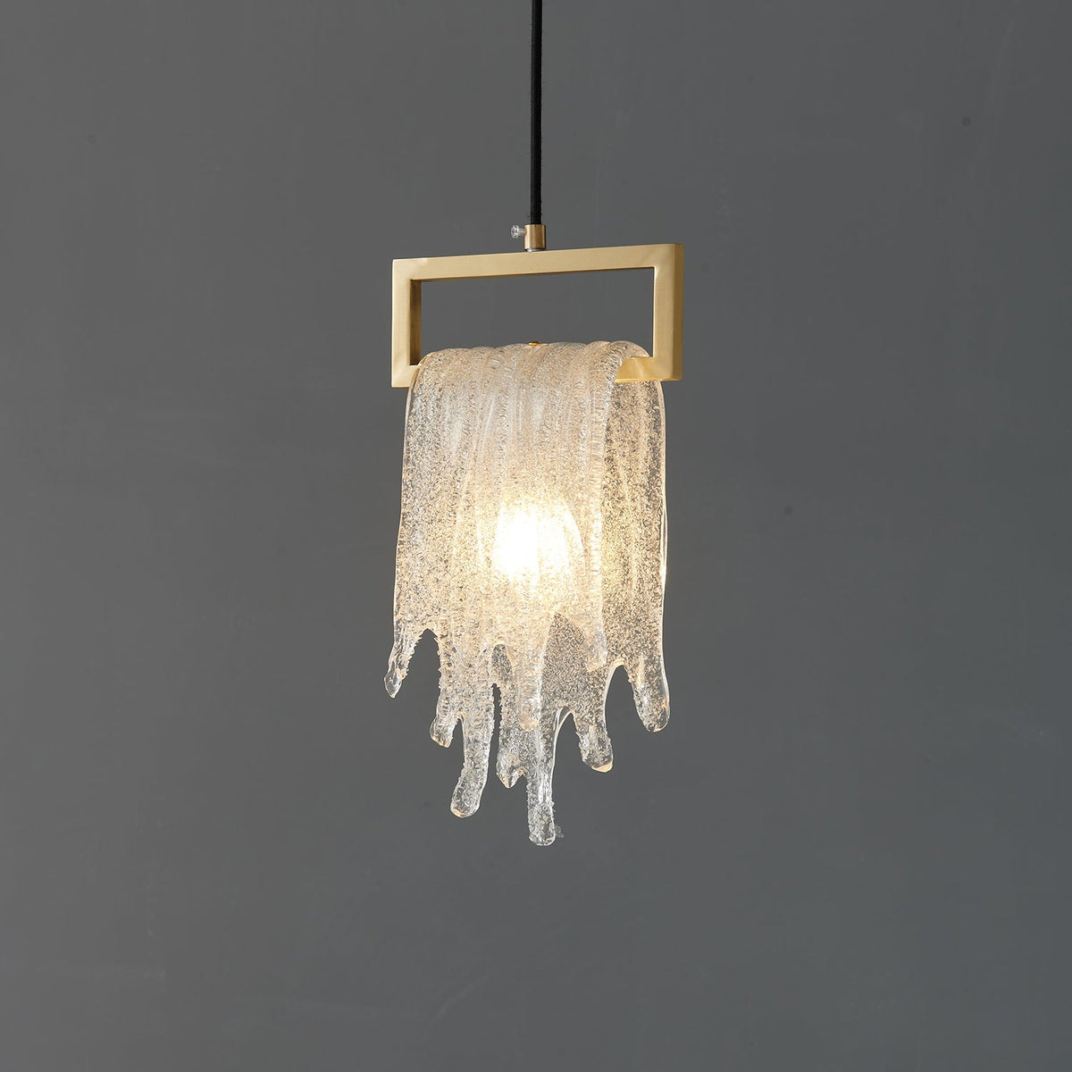 The same draped glass Clérachic pendant, lit, with the icicle-like glass glowing warm below a brass frame.