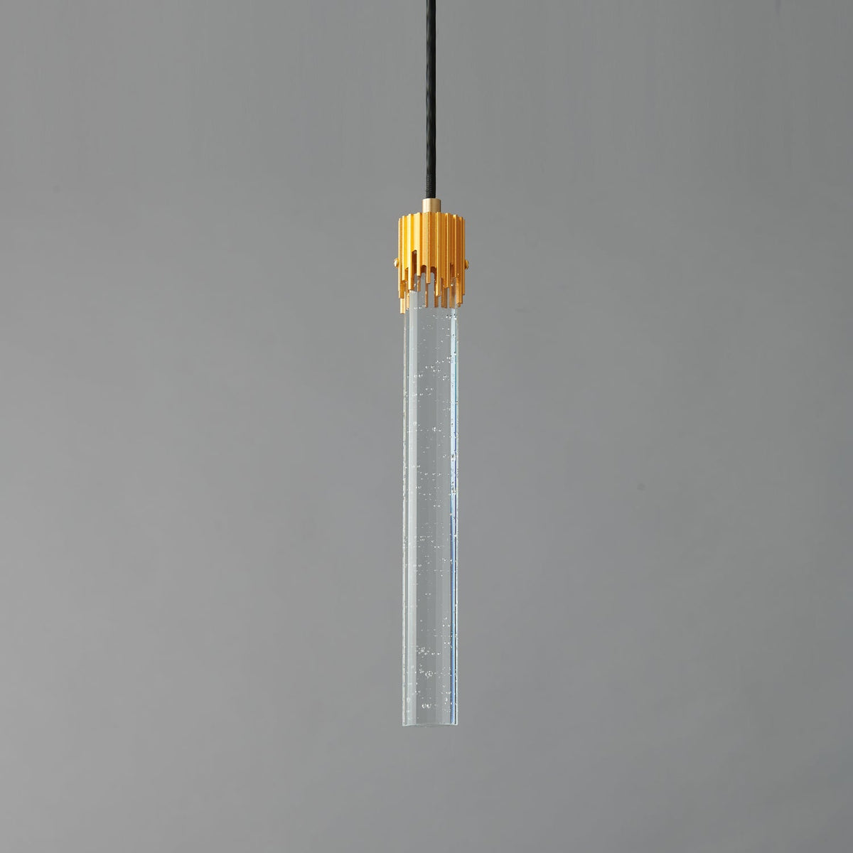A Clérachic pendant with a long clear glass rod flecked with tiny bubbles and a ribbed brass cap, unlit on grey.