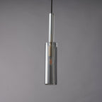 A Clérachic pendant with a slim smoke grey glass cylinder and a clear glass tube above it, unlit against a grey wall.