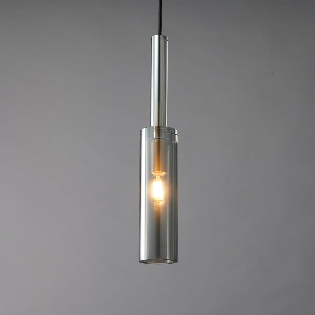 The smoke grey cylinder Clérachic pendant lit, with a warm filament bulb glowing inside the tinted glass.