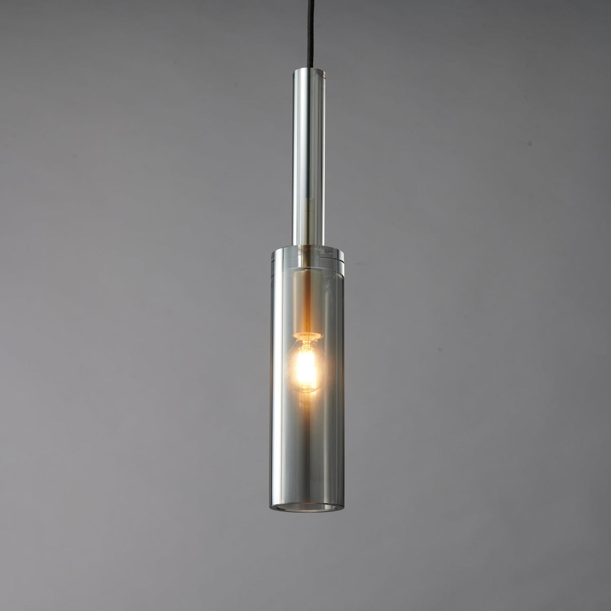 The smoke grey cylinder Clérachic pendant lit, with a warm filament bulb glowing inside the tinted glass.
