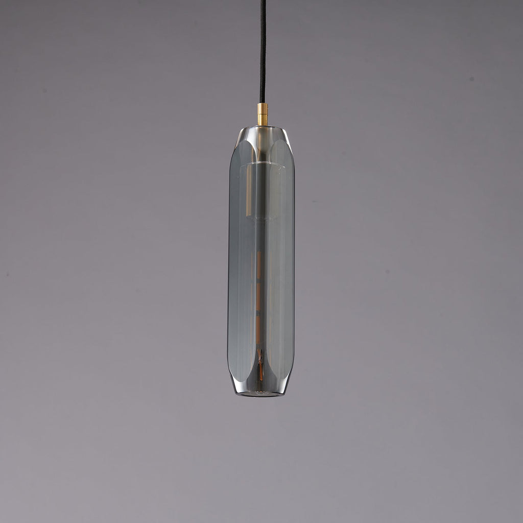 A tall, narrow Clérachic pendant with a smoke grey glass shade and a brass cap, unlit on a grey backdrop.
