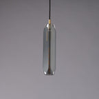 A tall, narrow Clérachic pendant with a smoke grey glass shade and a brass cap, unlit on a grey backdrop.