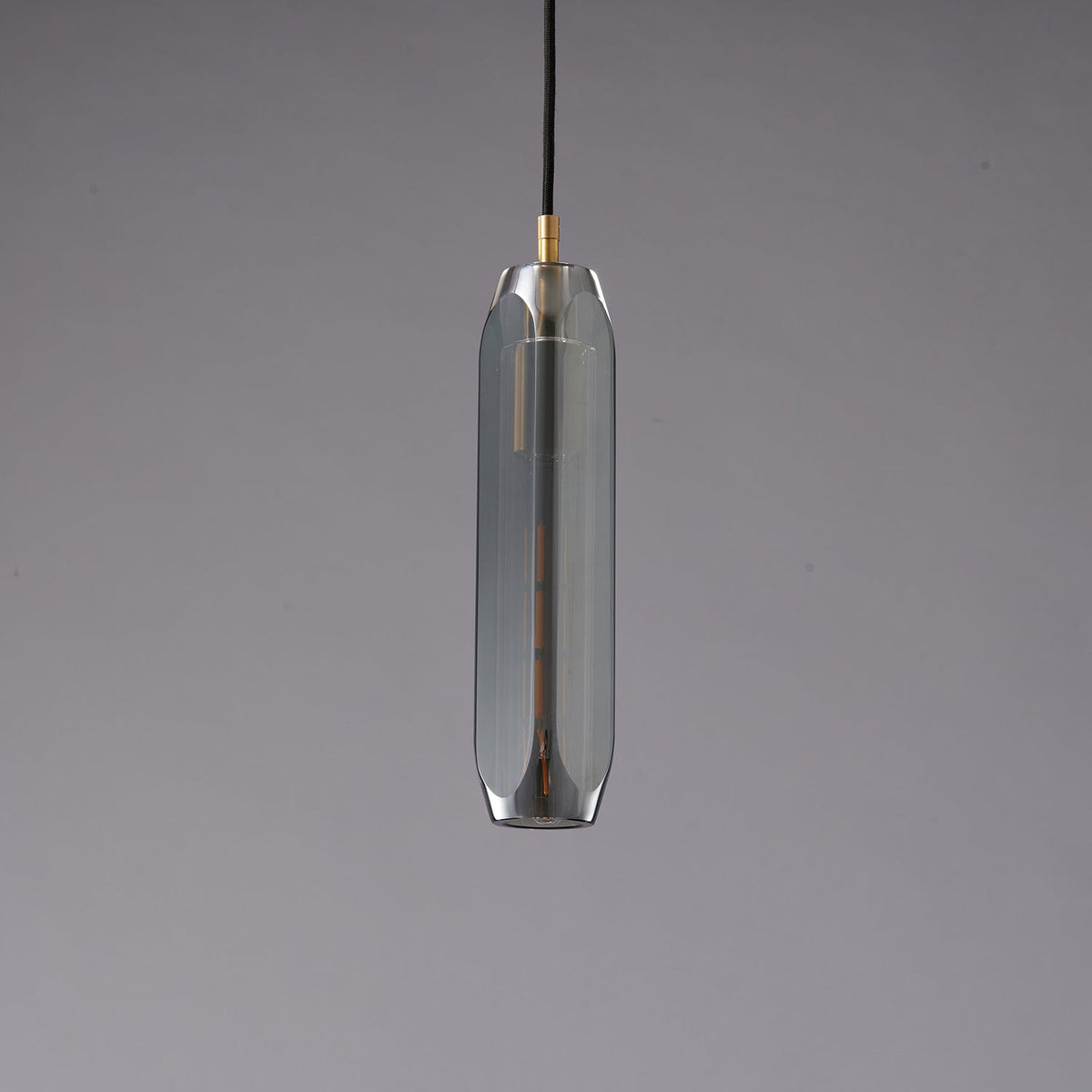 A tall, narrow Clérachic pendant with a smoke grey glass shade and a brass cap, unlit on a grey backdrop.