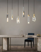 A portrait view of six Clérachic pendants in different glass shapes, hanging above a marble table and a navy chair.