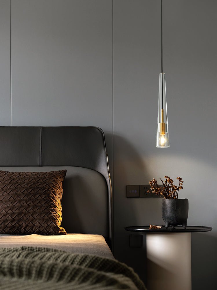 A tapered clear glass Clérachic pendant hangs by a leather headboard, in a portrait view with a dark side table.
