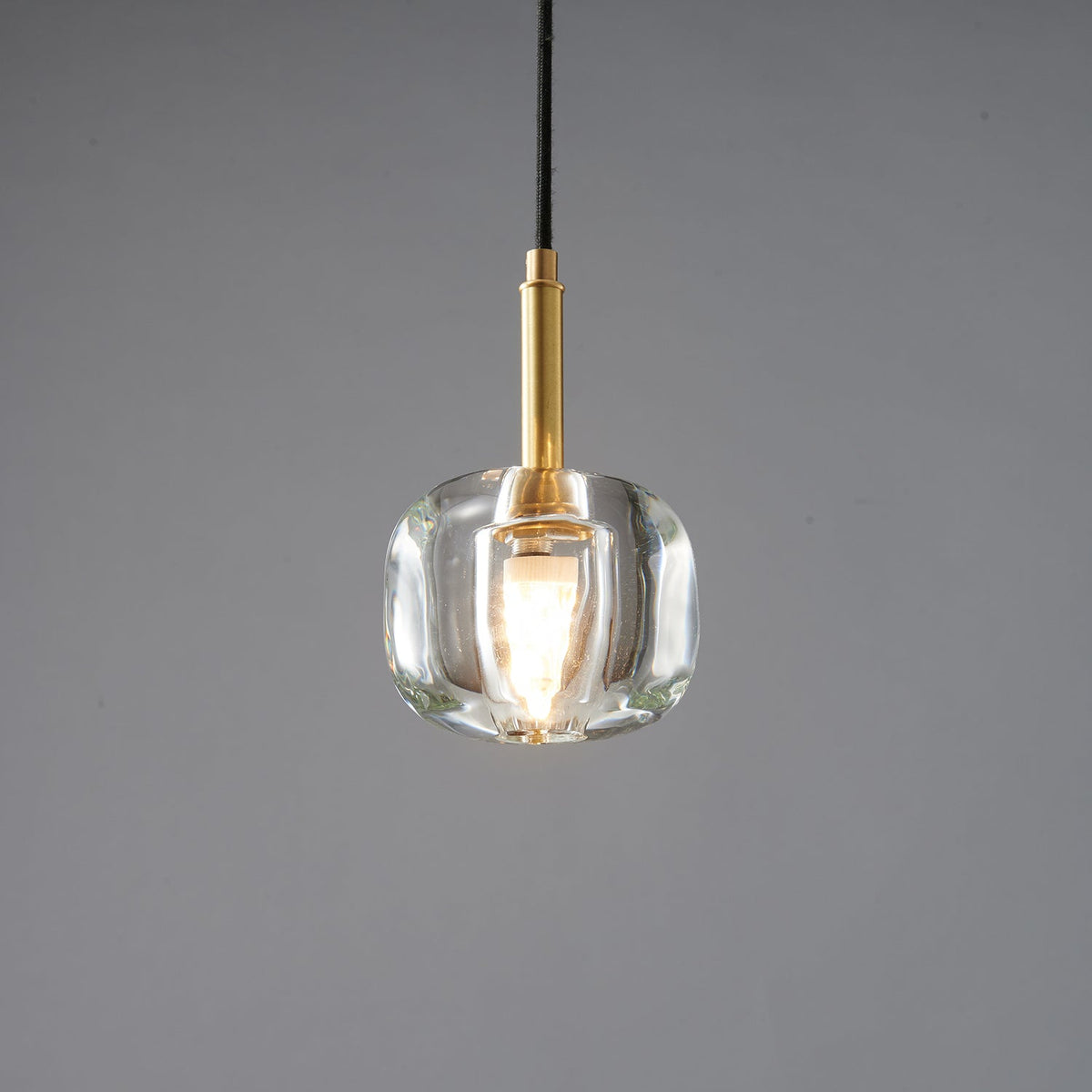 A Clérachic pendant with a rounded, thick clear glass shade and a brass stem, with a lit bulb inside, on a grey backdrop.