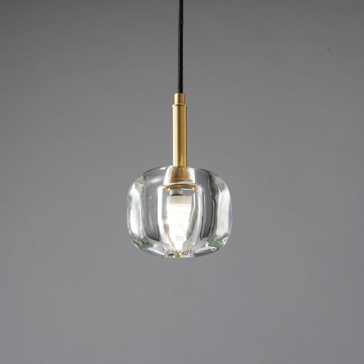 A smaller Clérachic pendant with a rounded, thick clear glass shade and a brass stem, lit on a grey backdrop.