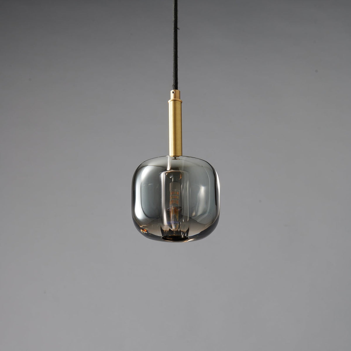 A Clérachic pendant with a rounded smoke grey glass shade and a brass stem, unlit on a grey backdrop.