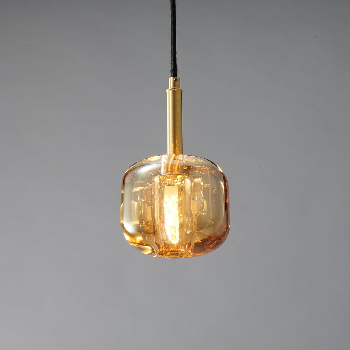A Clérachic pendant with a rounded amber glass shade and a brass stem, lit warm on a grey backdrop.
