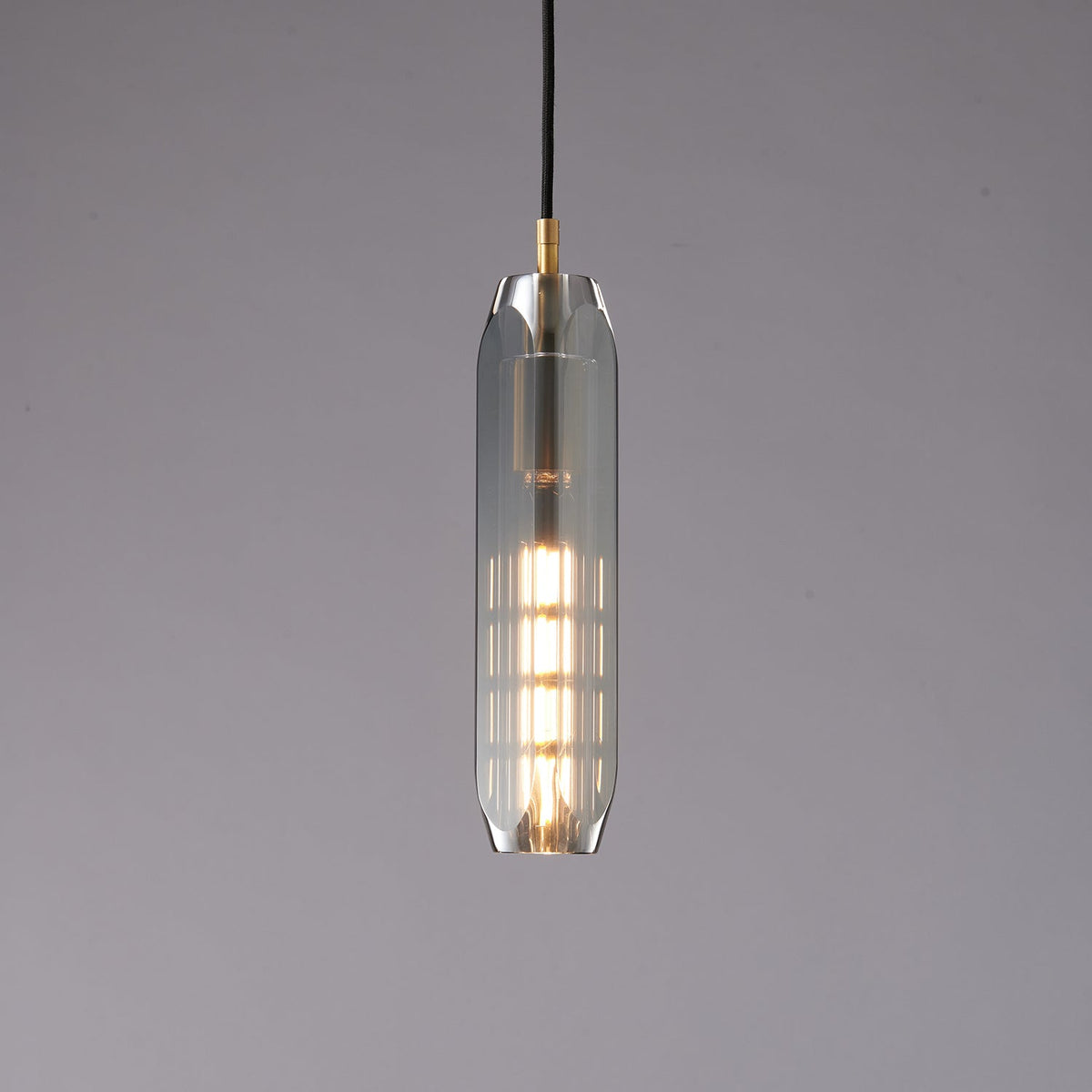 A tall, slim Clérachic pendant with a smoke grey glass shade, lit from within by a long filament bulb, on a grey backdrop.