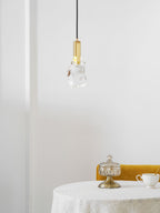 A Clérachic pendant with a rock-like clear glass piece and a brass bracket hangs above a round table with a glass jar.