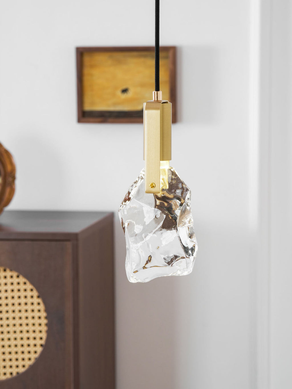 Rock-like clear glass on this Clérachic pendant, with a brass bracket, in front of a white panelled wall and a cabinet.