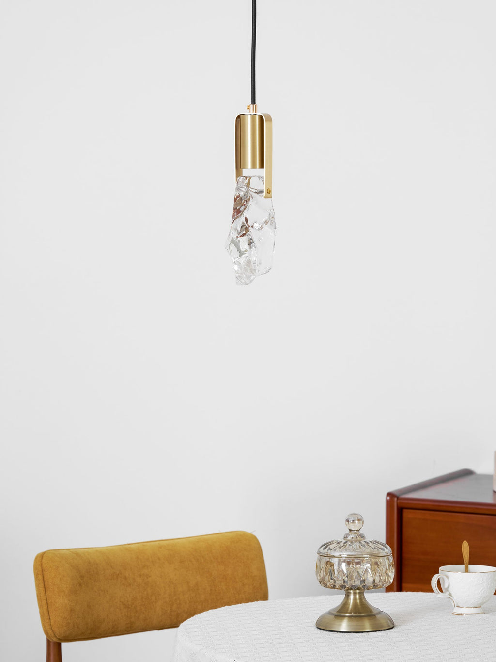 A small rock-glass Clérachic pendant with a round brass cap hangs above a table with a yellow chair and a cup.