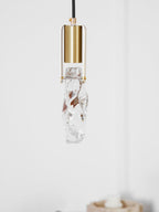 A Clérachic pendant with a long, uneven clear glass piece held in a brass cap with a thin frame, on a white wall.