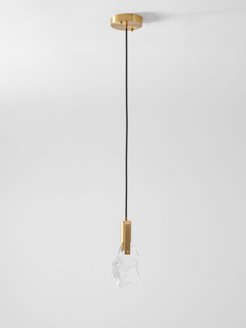 A single Clérachic pendant with a rock-like clear glass piece hangs from a brass ceiling canopy against a pale grey wall.