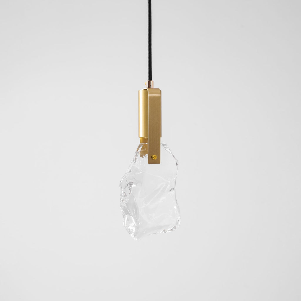 A Clérachic pendant with rough clear glass and a brass bracket, on a plain pale grey backdrop.