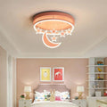 Clérachic round pink ceiling light with a glowing crescent moon and hanging stars above a bed in a soft pink child's room.