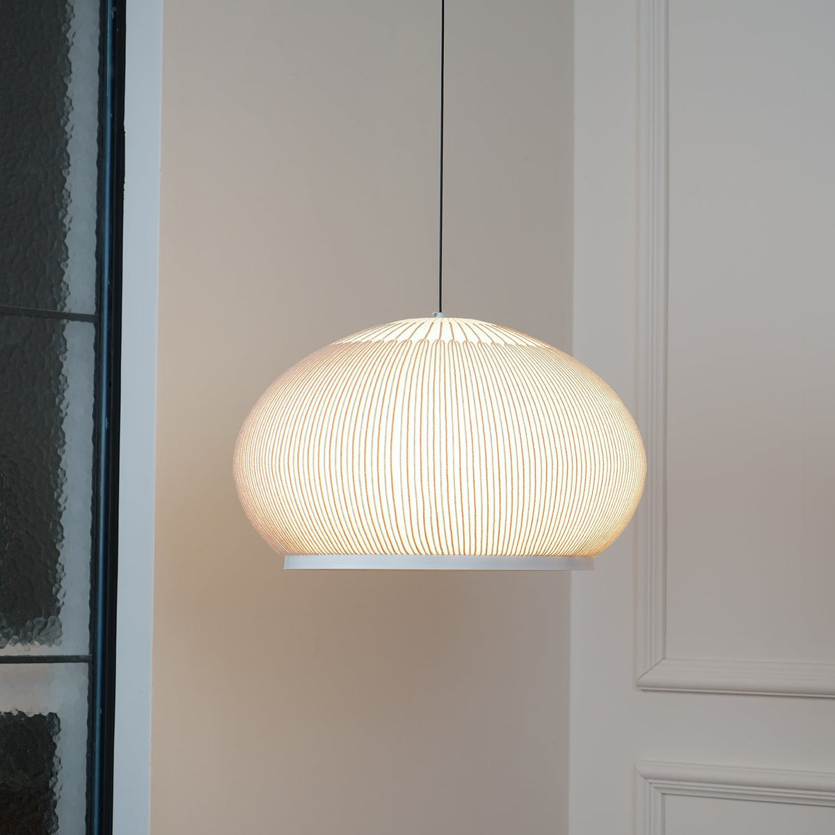 Clérachic wide dome knit pendant lamp glowing warm in a pink-toned corner, beside a tall black-framed glass door.