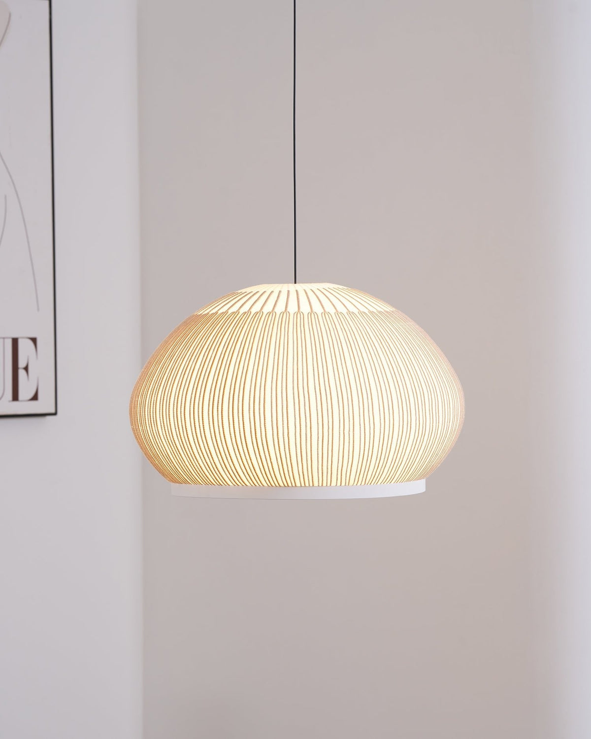 Clérachic wide dome knit pendant lamp lit warm in daylight, hung close against the pale corner wall beside the framed print.