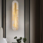 Clérachic Kristy wall lamp lit above a bed with a monstera leaf in a glass vase.