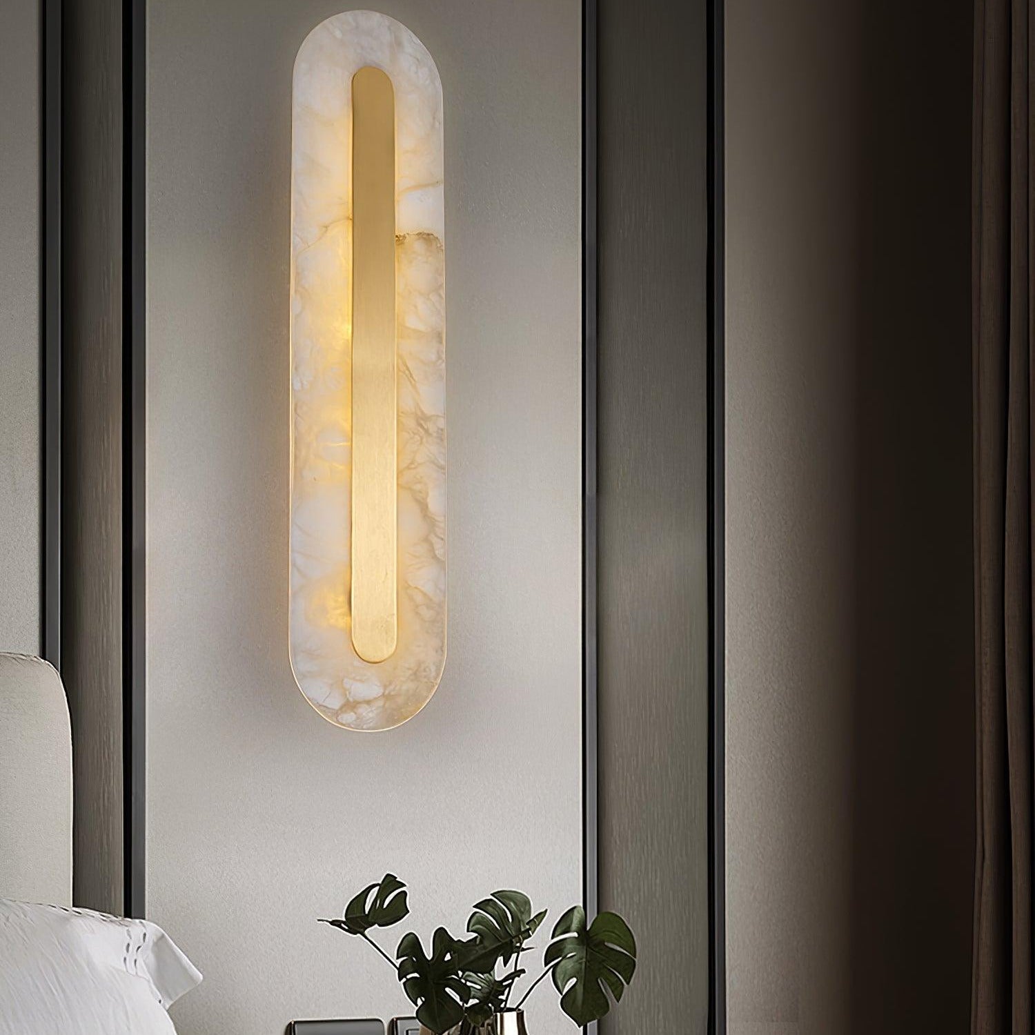 Clérachic Kristy wall lamp lit above a bed with a monstera leaf in a glass vase.