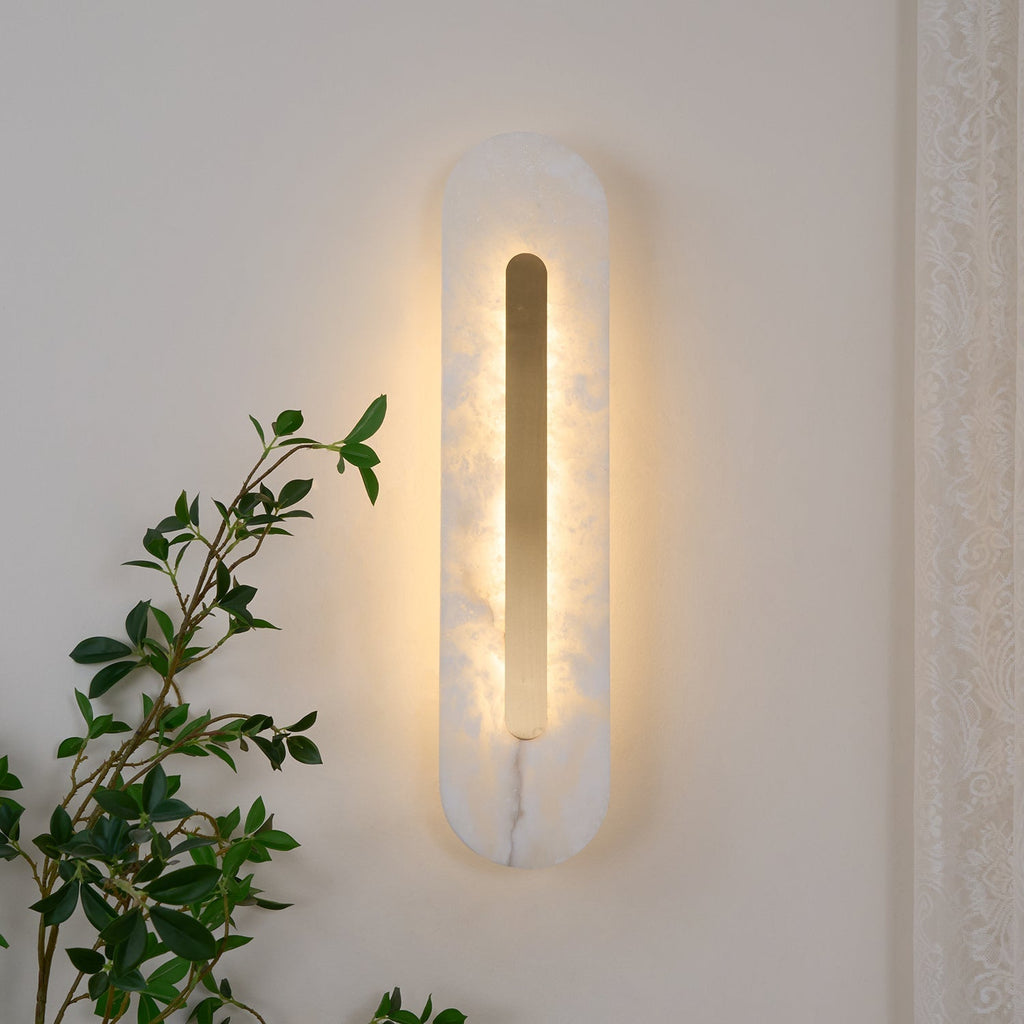 Clérachic Kristy wall lamp lit on cream plaster with green foliage trailing nearby.