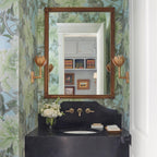 Clérachic brass tulip sconces flank a gilt mirror above a black stone vanity in a floral-papered bath.