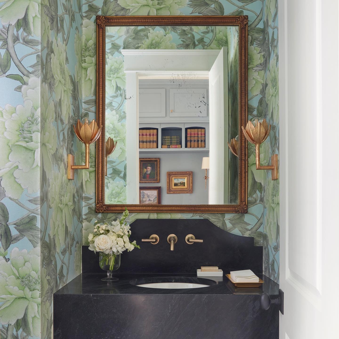 Clérachic brass tulip sconces flank a gilt mirror above a black stone vanity in a floral-papered bath.