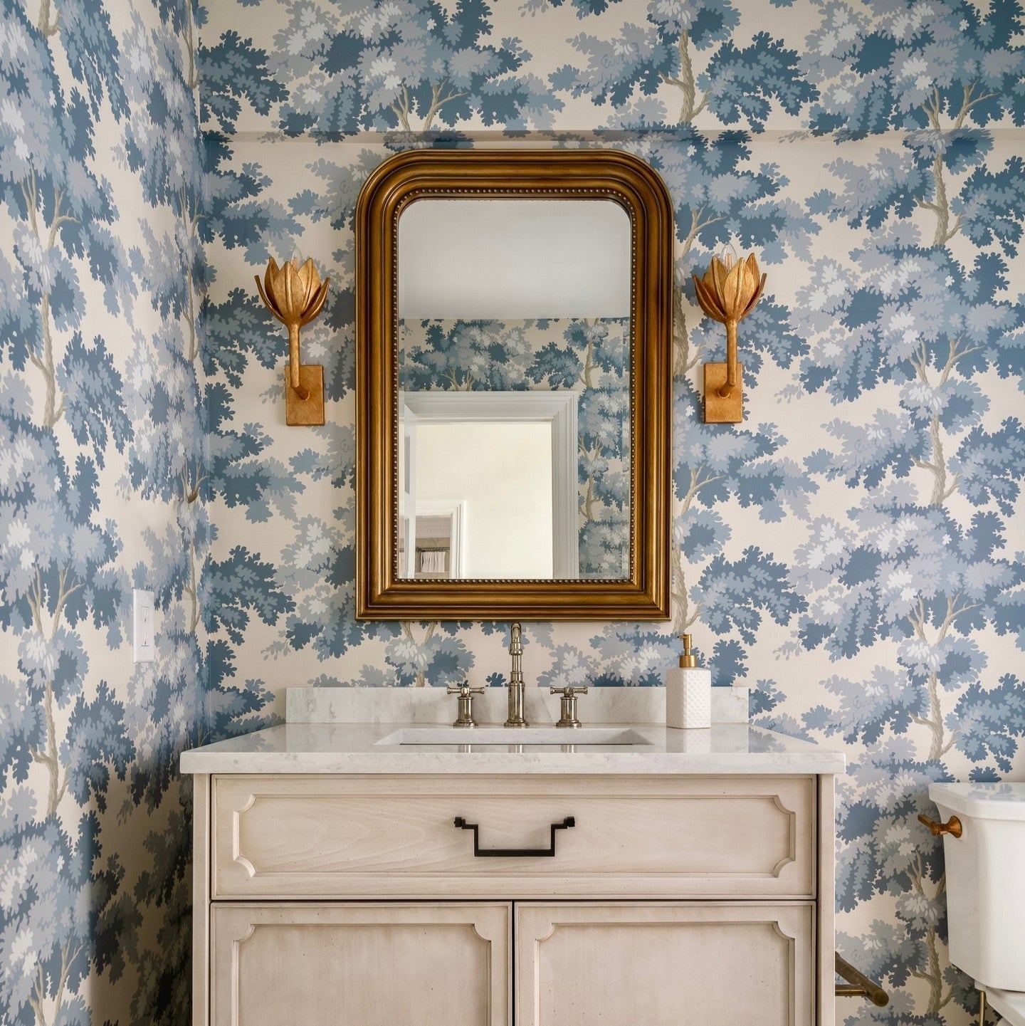 The Clérachic sconces glow beside a gold mirror above a pale wood vanity in a blue floral powder room.
