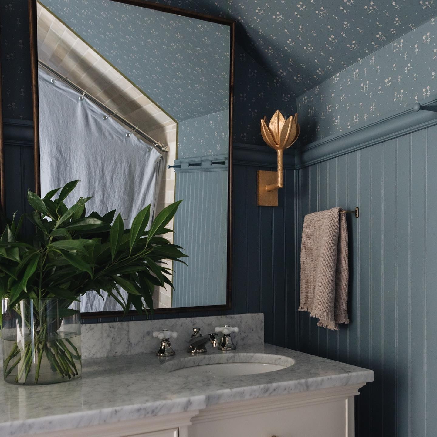 A Clérachic brass sconce lights a black-framed mirror in a moody teal-paneled bathroom.