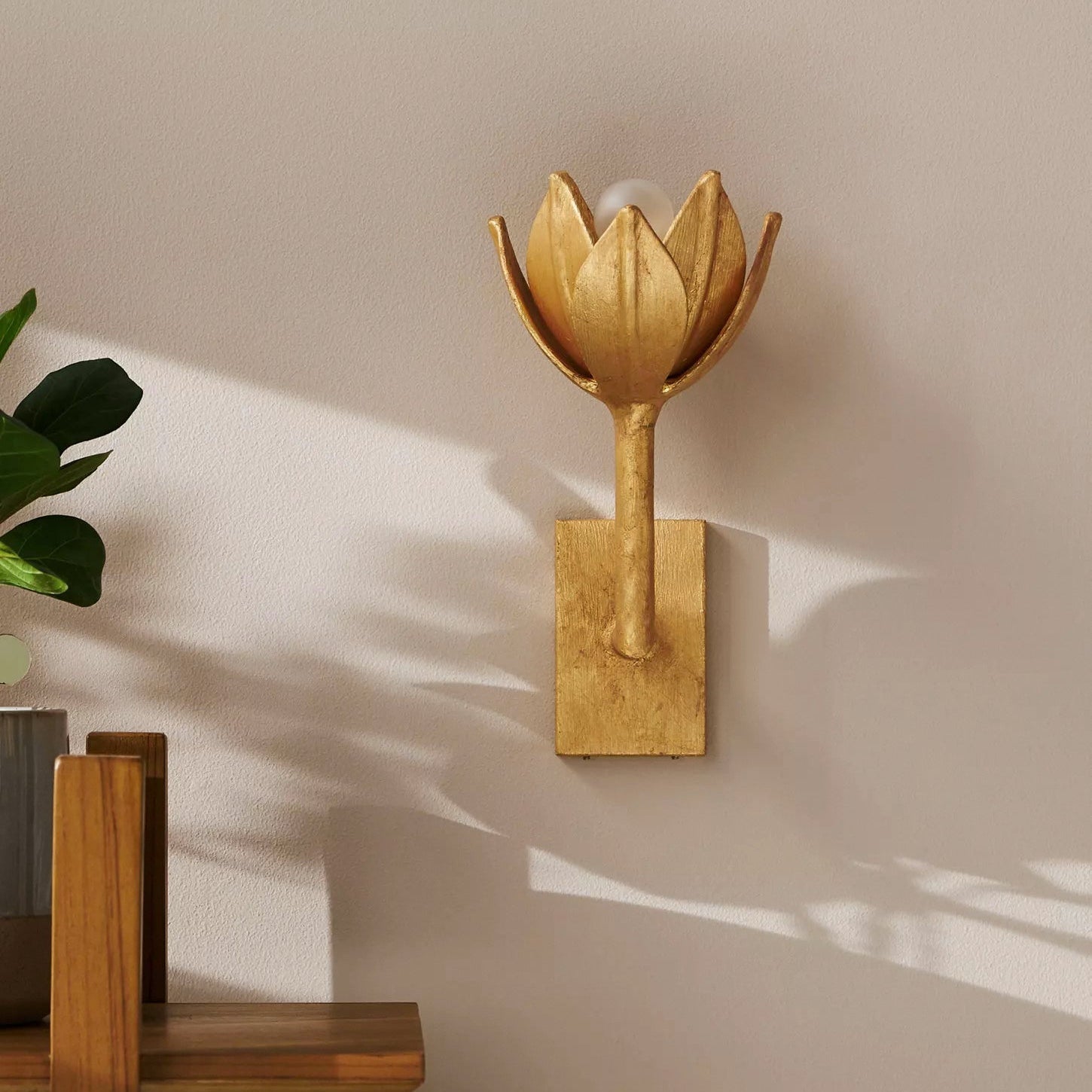 A Clérachic brass tulip sconce mounts on a soft pink wall beside a leafy potted plant.