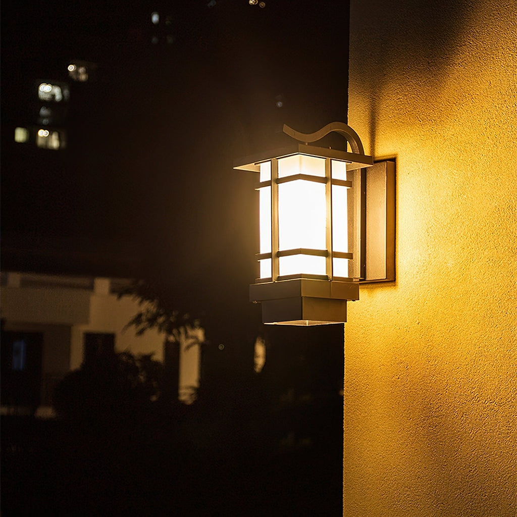 Lit Clérachic black lantern wall light on a pale exterior wall at night, its frosted glass glowing warm yellow.