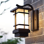 Clérachic lantern wall light on a grey brick wall, its black frame and frosted panels lit, with greenery blurred below.