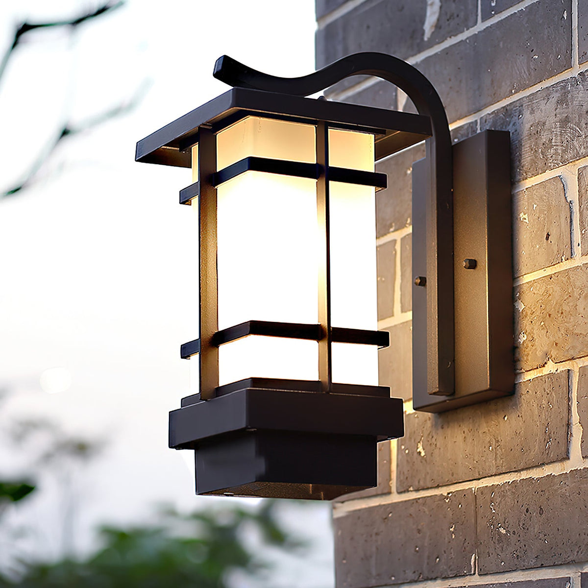 Clérachic lantern wall light on a grey brick wall, its black frame and frosted panels lit, with greenery blurred below.