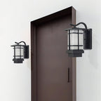 Two Clérachic lantern wall lights flank a dark brown door on a white wall, one smaller than the other, both unlit.