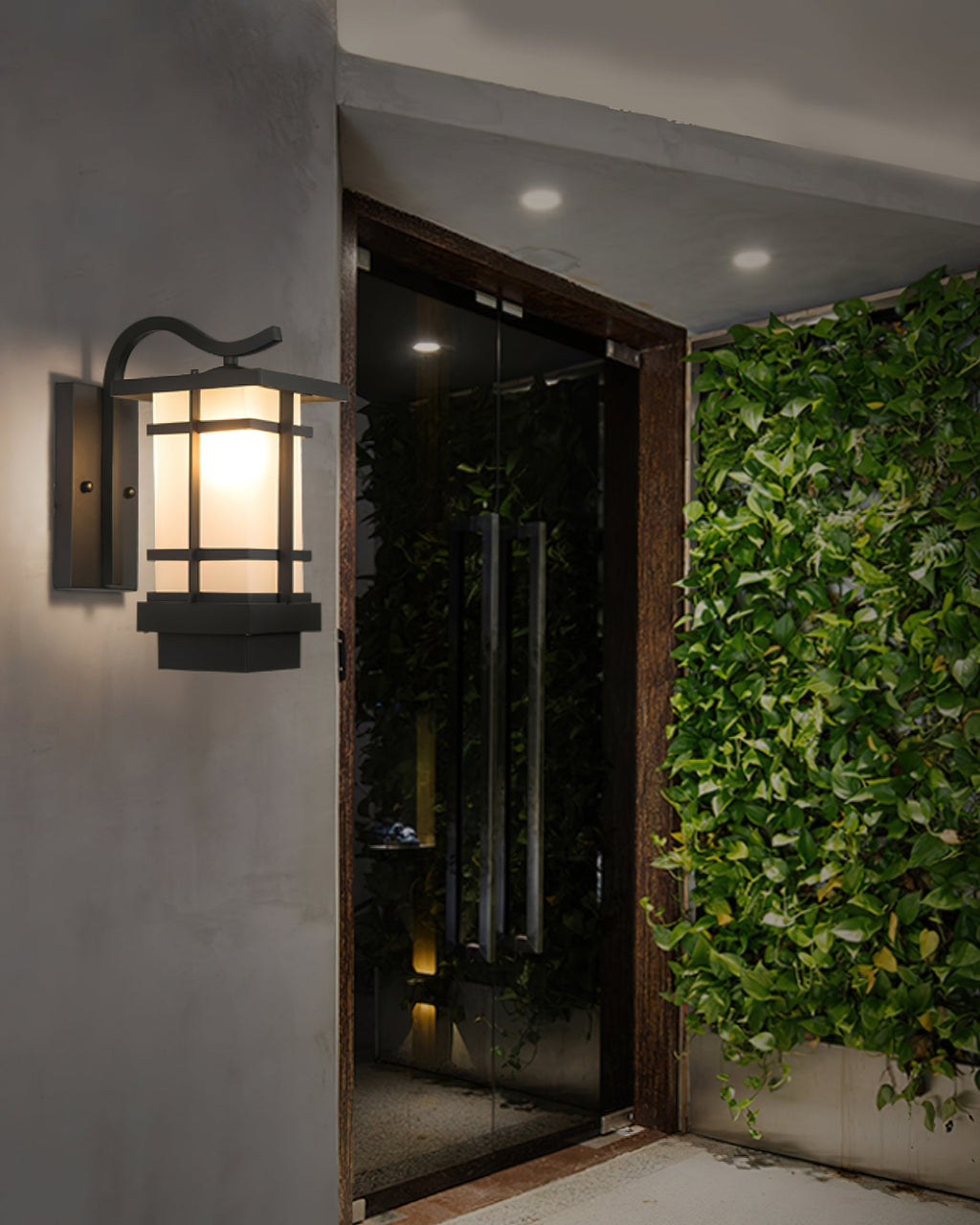 A Clérachic lantern wall light lit beside a dark glass entrance, with a wall of green ivy to the right.