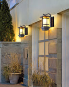 A pair of lit Clérachic lantern wall lights on a stone-clad entrance wall at dusk, with potted grasses below.