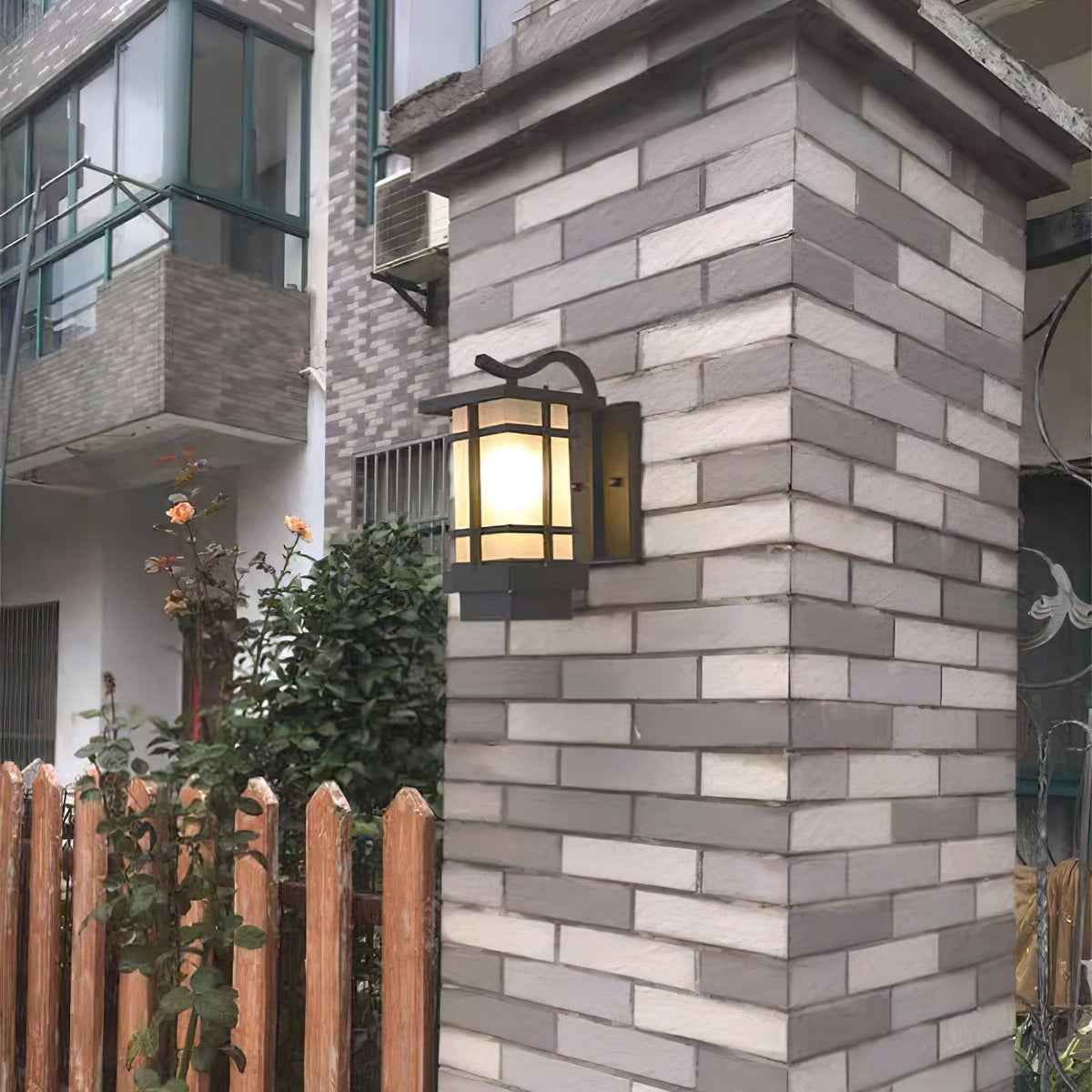 Lit Clérachic lantern wall light on a grey brick gatepost, a wooden fence and a rose bush beside it.