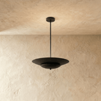 Clérachic pendant light in matte black, a wide disc shade hanging from a slim rod against a plaster wall.