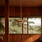 Clérachic pendant light in brushed brass suspended near a window overlooking trees and the coastline.