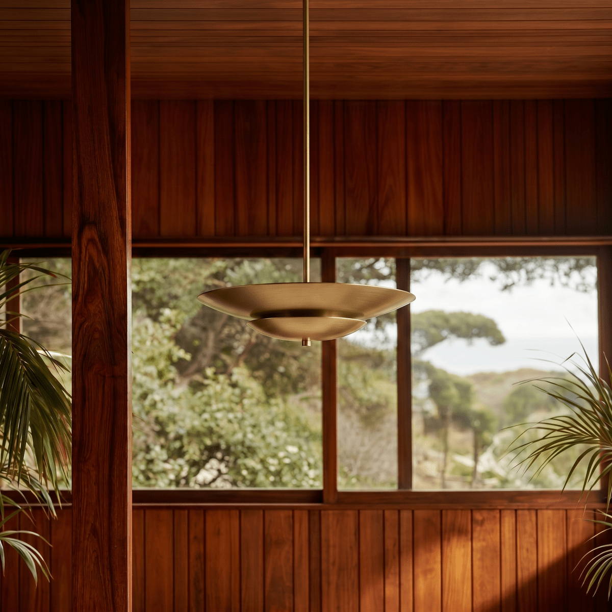 Clérachic pendant light in brushed brass suspended near a window overlooking trees and the coastline.