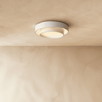 Clérachic white layered round flush ceiling light seen from below, its inner drum lit warmly against a plastered ceiling.