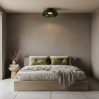 A green glass Clérachic ceiling light above a low platform bed with olive cushions, against a textured taupe wall.