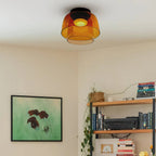 An orange glass Clérachic ceiling light in the corner of a cream room with a framed print and a loaded bookshelf.