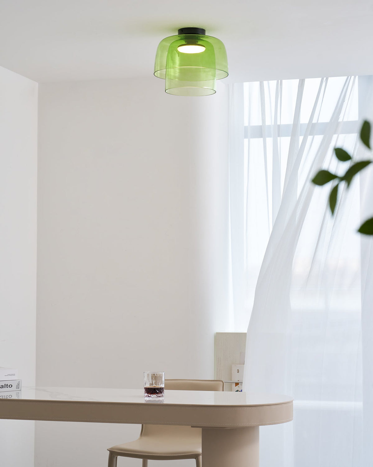 A green glass Clérachic ceiling light, lit, above a narrow table with a glass of dark drink and a leafy branch to the right.