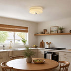 Lit layered glass Clérachic flush mount above a round wooden table in a kitchen with a bamboo blind and an open shelf.